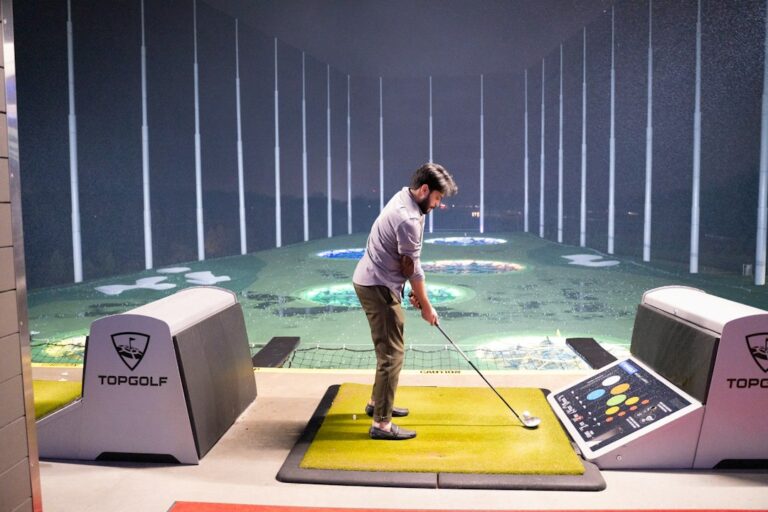 Golfer checking alignment beside a simulator screen while practicing an iron shot from a hitting mat.